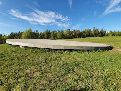 A large circular concrete structure is situated on a grassy landscape. In the background, there are trees under a blue sky with scattered white clouds. The structure appears to be slightly raised above the ground.
