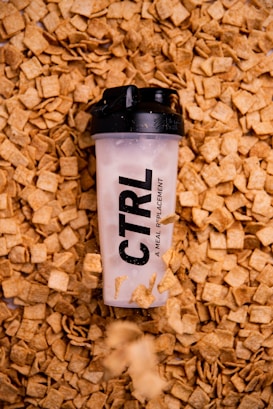 A meal replacement shaker bottle with a black lid is lying on a bed of square-shaped cereal pieces. The bottle has the word 'CTRL' printed on it, and some cereal pieces appear to be floating or falling around it.