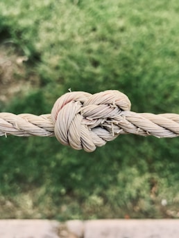 A tightly tied knot made of natural fiber rope against a blurred, green grass background. The texture of the rope is rough and fibrous, with visible strands.