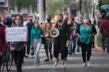 A group of people wearing face masks are walking down a city street. One person at the front is speaking into a megaphone, leading the march. Another person holds a sign with a message advocating for the value of a Black child's life. The crowd appears to be engaged in a protest or demonstration.