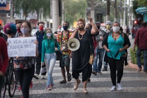 A group of people wearing face masks are walking down a city street. One person at the front is speaking into a megaphone, leading the march. Another person holds a sign with a message advocating for the value of a Black child's life. The crowd appears to be engaged in a protest or demonstration.