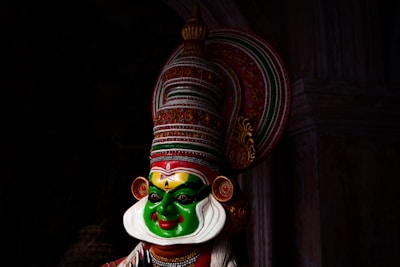 A vibrant and colorful traditional mask featuring intricate designs and patterns. The mask's face is predominantly green with white and red details, including a large headdress adorned with elaborate ornamentation. The expression appears dramatic, characteristic of classical dance forms.