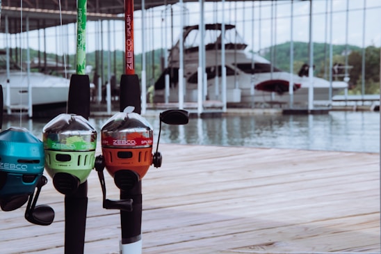 Several brightly colored fishing rods with Zebco Splash branding are mounted on a wooden dock. In the background, a large boat is docked in a marina, with green hills visible across the water.