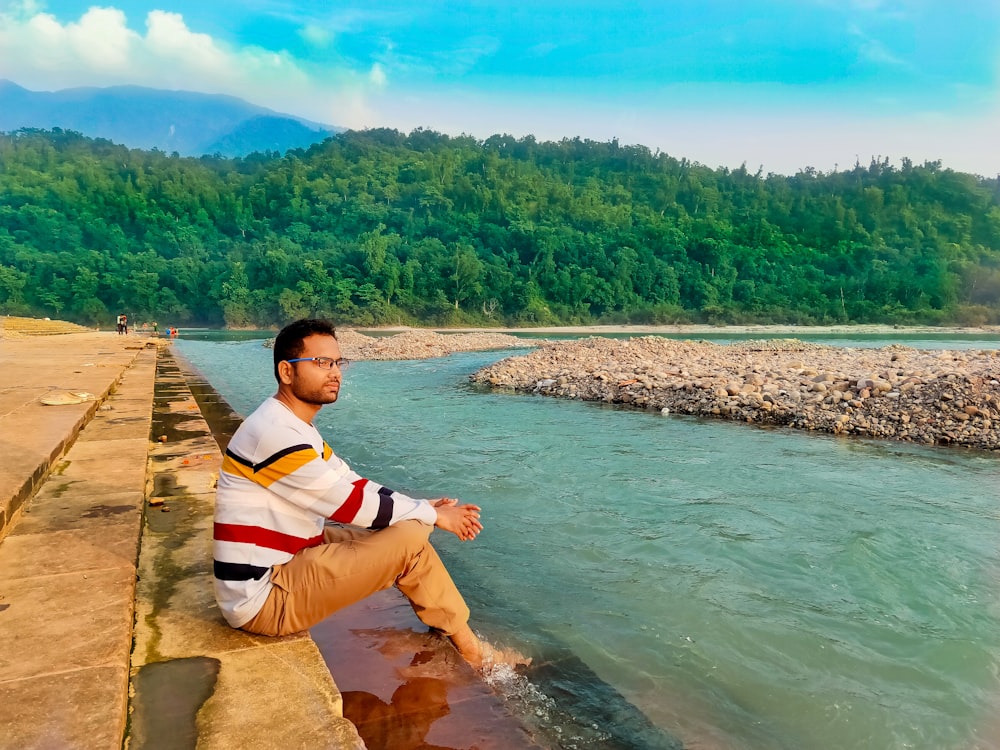 man in white and blue striped polo shirt sitting on brown wooden dock during daytime