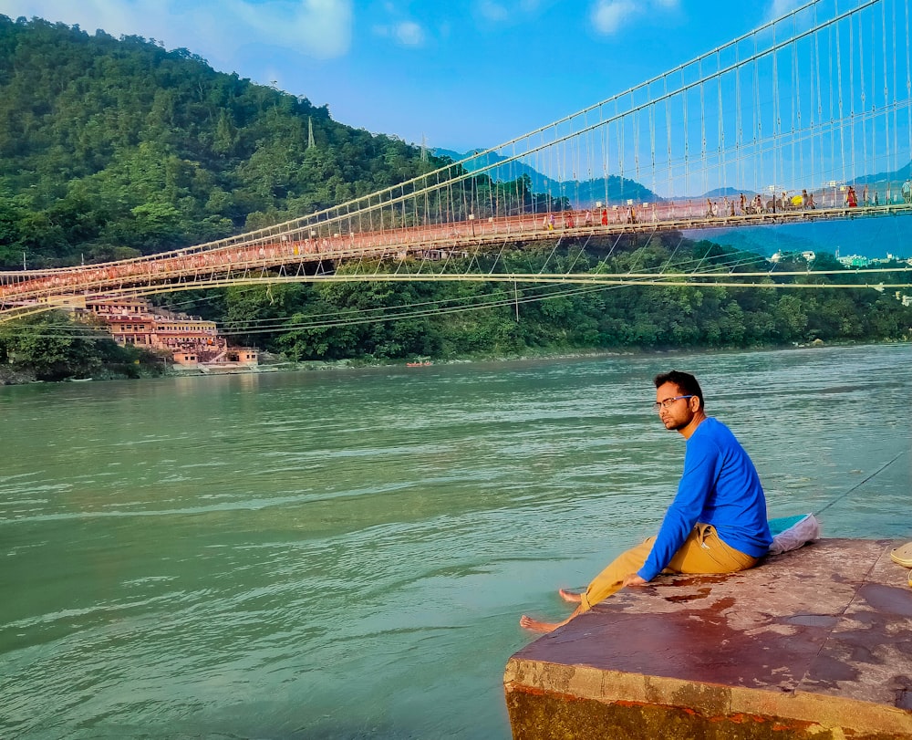 man in blue shirt sitting on brown rock near body of water during daytime