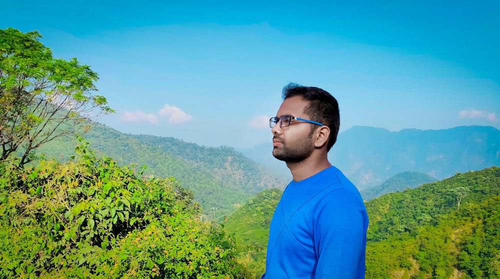 man in blue crew neck shirt wearing black framed eyeglasses standing near green plants during daytime