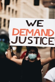 A person wearing a face mask and glasses holds up a sign that reads 'We Demand Justice' during a protest. The background is blurred, showing other participants and city buildings.