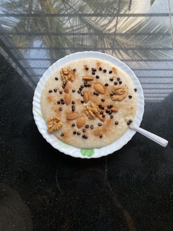 A bowl of oatmeal topped with various nuts and small chocolate chips. The toppings include walnuts and almonds, arranged neatly across the surface. The bowl is placed on a reflective dark countertop, with natural light coming in from the surrounding area.