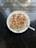 A bowl of oatmeal topped with various nuts and small chocolate chips. The toppings include walnuts and almonds, arranged neatly across the surface. The bowl is placed on a reflective dark countertop, with natural light coming in from the surrounding area.