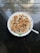 A bowl of oatmeal topped with various nuts and small chocolate chips. The toppings include walnuts and almonds, arranged neatly across the surface. The bowl is placed on a reflective dark countertop, with natural light coming in from the surrounding area.