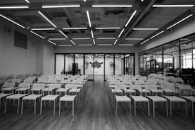 An empty conference room with multiple rows of white chairs arranged in a neat grid pattern on a wooden floor. The ceiling features exposed ducts and long, linear lighting fixtures. A glass wall at the far end reveals an adjacent workspace with colorful decor visible through the transparent panels.