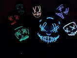 Several glowing masks with eerie, stitched-smile designs and crossed eyes are illuminated in various colors against a dark background, creating a mysterious and unsettling atmosphere.
