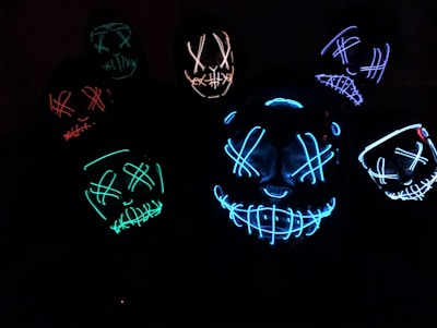 Several glowing masks with eerie, stitched-smile designs and crossed eyes are illuminated in various colors against a dark background, creating a mysterious and unsettling atmosphere.