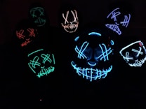 Several glowing masks with eerie, stitched-smile designs and crossed eyes are illuminated in various colors against a dark background, creating a mysterious and unsettling atmosphere.