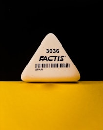 A triangular white eraser with the brand name 'FACTIS' and the number '3036' printed on it, along with a barcode and the word 'SPAIN'. The eraser is positioned against a contrasting black and yellow background.