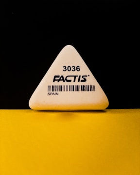 A triangular white eraser with the brand name 'FACTIS' and the number '3036' printed on it, along with a barcode and the word 'SPAIN'. The eraser is positioned against a contrasting black and yellow background.