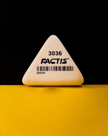 A triangular white eraser with the brand name 'FACTIS' and the number '3036' printed on it, along with a barcode and the word 'SPAIN'. The eraser is positioned against a contrasting black and yellow background.