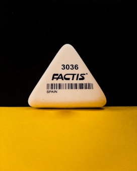 A triangular white eraser with the brand name 'FACTIS' and the number '3036' printed on it, along with a barcode and the word 'SPAIN'. The eraser is positioned against a contrasting black and yellow background.