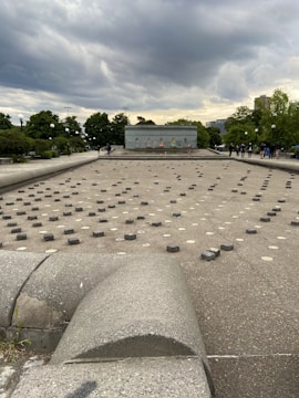A large concrete area features numerous small stone pavers arranged in rows on the ground. In the background, a low wall displays colorful graffiti. The scene is set under a cloudy sky with visible storm clouds, surrounded by green trees and a few people walking or standing around.