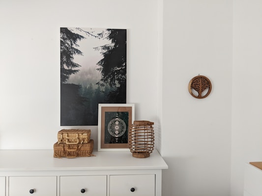 A white cabinet with black handles is topped with various decorative items including a wicker basket, framed artwork, and a lantern. Above the cabinet, a photograph of a misty forest is hung on the wall. There is also a wooden wall decoration featuring a tree motif to the right of the photograph.