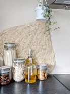 A modern kitchen setup featuring glass jars filled with dried ingredients like pasta, oats, dates, and cookies. A glass bottle containing a yellow liquid, possibly oil, stands in the middle. A woven basket decoration is placed on the wall, and a hanging plant in a white pot adds a touch of greenery.