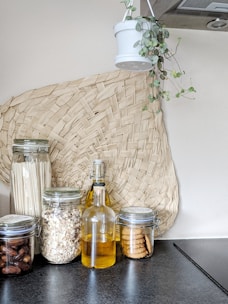 A modern kitchen setup featuring glass jars filled with dried ingredients like pasta, oats, dates, and cookies. A glass bottle containing a yellow liquid, possibly oil, stands in the middle. A woven basket decoration is placed on the wall, and a hanging plant in a white pot adds a touch of greenery.