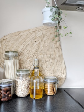 A modern kitchen setup featuring glass jars filled with dried ingredients like pasta, oats, dates, and cookies. A glass bottle containing a yellow liquid, possibly oil, stands in the middle. A woven basket decoration is placed on the wall, and a hanging plant in a white pot adds a touch of greenery.