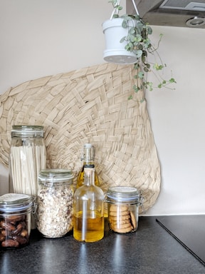 A modern kitchen setup featuring glass jars filled with dried ingredients like pasta, oats, dates, and cookies. A glass bottle containing a yellow liquid, possibly oil, stands in the middle. A woven basket decoration is placed on the wall, and a hanging plant in a white pot adds a touch of greenery.