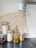 A modern kitchen setup featuring glass jars filled with dried ingredients like pasta, oats, dates, and cookies. A glass bottle containing a yellow liquid, possibly oil, stands in the middle. A woven basket decoration is placed on the wall, and a hanging plant in a white pot adds a touch of greenery.
