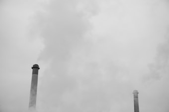 Two large industrial smokestacks emit smoke into an overcast sky, blending into the cloudy, gray atmosphere around them.