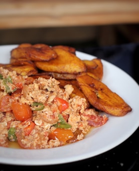 A white plate holds a serving of fried plantains accompanied by a colorful scrambled eggs mixture that includes chopped green peppers, tomatoes, and onions.