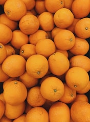 A pile of fresh oranges with a smooth texture and vibrant color, closely packed together. The surface of each orange displays slight variations with small dimples and natural imperfections.