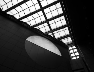 Architectural structure with a geometric design featuring a large grid-patterned glass ceiling and a circular window on the wall. The scene is shown in black and white, emphasizing the contrast between light and shadow.