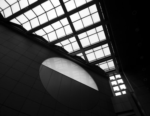 Architectural structure with a geometric design featuring a large grid-patterned glass ceiling and a circular window on the wall. The scene is shown in black and white, emphasizing the contrast between light and shadow.