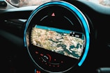 A circular car infotainment display features a GPS navigation interface showing a detailed map with various geographic features. The interface includes navigation icons and options beneath the display, and the outer rim is illuminated with a blue LED light.