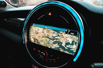 A circular car infotainment display features a GPS navigation interface showing a detailed map with various geographic features. The interface includes navigation icons and options beneath the display, and the outer rim is illuminated with a blue LED light.