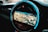 A circular car infotainment display features a GPS navigation interface showing a detailed map with various geographic features. The interface includes navigation icons and options beneath the display, and the outer rim is illuminated with a blue LED light.
