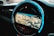 A circular car infotainment display features a GPS navigation interface showing a detailed map with various geographic features. The interface includes navigation icons and options beneath the display, and the outer rim is illuminated with a blue LED light.