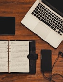 A wooden desk setup featuring a laptop with a silver body and black keyboard, an open notebook with handwritten notes, a smartphone with earphones plugged in, and a black tablet.