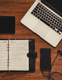 A wooden desk setup featuring a laptop with a silver body and black keyboard, an open notebook with handwritten notes, a smartphone with earphones plugged in, and a black tablet.