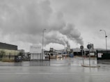 A factory environment with large amounts of smoke billowing from chimneys under an overcast sky. The foreground features an industrial entrance with a sign, gate, and street lamps. A truck is visible near the center, and the ground is wet, indicating recent rain.