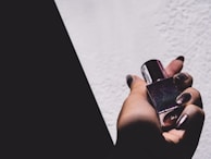 A hand with polished nails holds a bottle of nail polish against a contrasting background, creating a dramatic shadow effect. The lighting highlights the reflective surface of the nail polish.