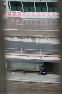 An urban setting with wet pavement and a person seated on a sidewalk corner beneath a modern building. The individual appears to be bundled in clothing with bags beside them, likely suggesting they are resting or taking shelter. In the background, there is a reflective glass facade with a red and white construction barrier separating the building from the street.