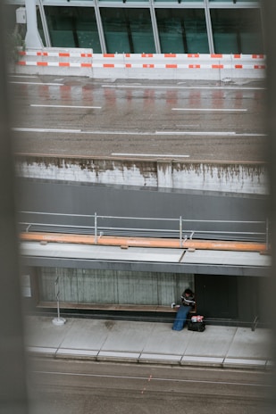 An urban setting with wet pavement and a person seated on a sidewalk corner beneath a modern building. The individual appears to be bundled in clothing with bags beside them, likely suggesting they are resting or taking shelter. In the background, there is a reflective glass facade with a red and white construction barrier separating the building from the street.