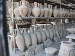 A collection of ancient pottery and ceramic artifacts, including tall amphorae, are arranged on metal shelves. These artifacts appear to be part of an archaeological storage or display, set against a backdrop of what seems to be stone walls.