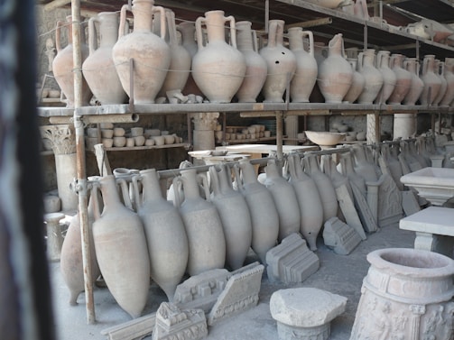 A collection of ancient pottery and ceramic artifacts, including tall amphorae, are arranged on metal shelves. These artifacts appear to be part of an archaeological storage or display, set against a backdrop of what seems to be stone walls.