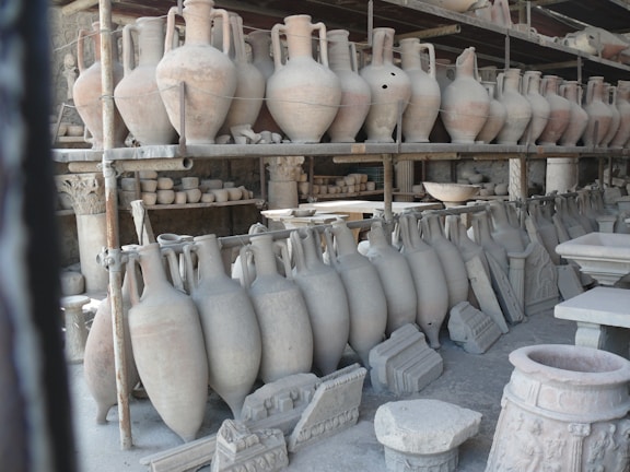 A collection of ancient pottery and ceramic artifacts, including tall amphorae, are arranged on metal shelves. These artifacts appear to be part of an archaeological storage or display, set against a backdrop of what seems to be stone walls.