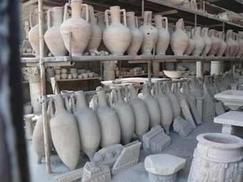 A collection of ancient pottery and ceramic artifacts, including tall amphorae, are arranged on metal shelves. These artifacts appear to be part of an archaeological storage or display, set against a backdrop of what seems to be stone walls.