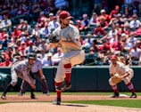 A baseball pitcher is in the middle of his windup on a sunny day during a game. He is wearing a uniform with red accents and a cap, and there is a crowd in the stands, most likely fans, watching intently. An opposing player is positioned nearby, possibly getting ready to run.