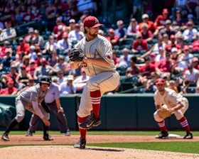 A baseball pitcher is in the middle of his windup on a sunny day during a game. He is wearing a uniform with red accents and a cap, and there is a crowd in the stands, most likely fans, watching intently. An opposing player is positioned nearby, possibly getting ready to run.